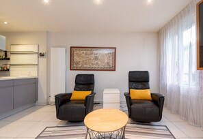 Ein modernes Wohnzimmer mit zwei schwarzen Ledersesseln, einem Holztisch, einer Küchenzeile mit weißer Arbeitsplatte und einem großen gerahmten Kunstwerk an der Wand.