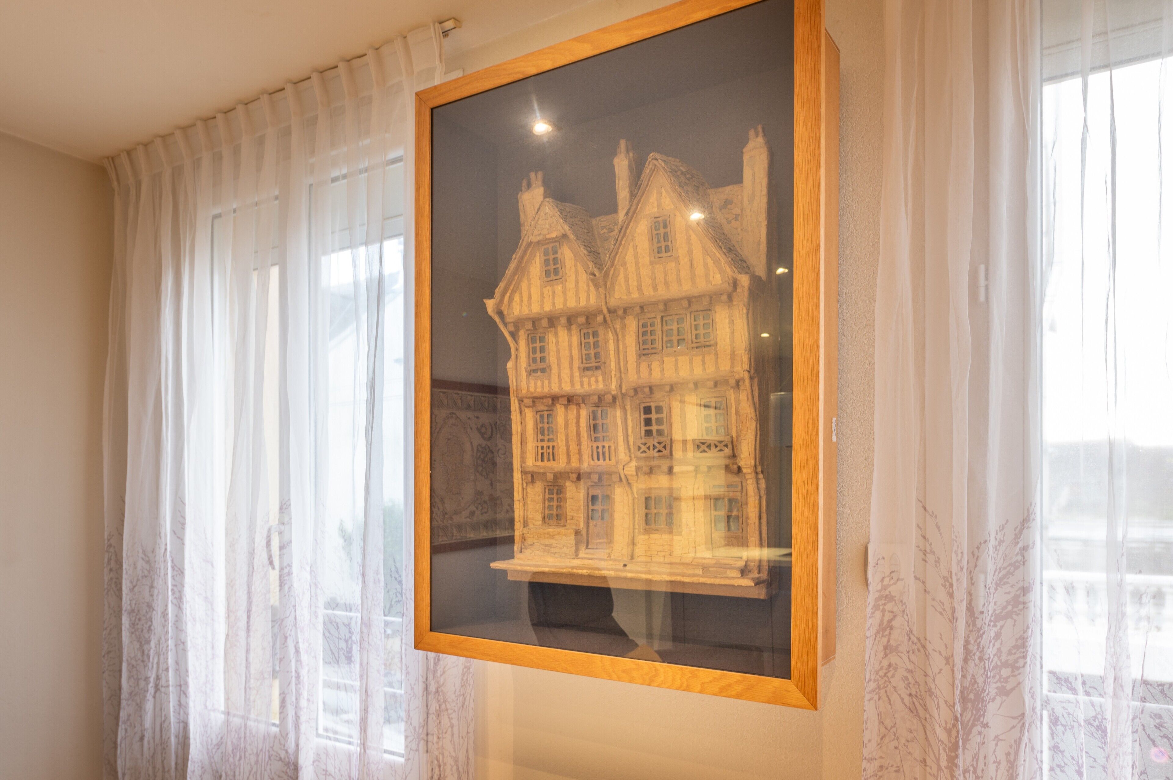 Model arquitectònic emmarcat d'un edifici amb diverses finestres i xemeneies, exposat en una habitació amb cortines lleugeres i una finestra que mostra un balcó.