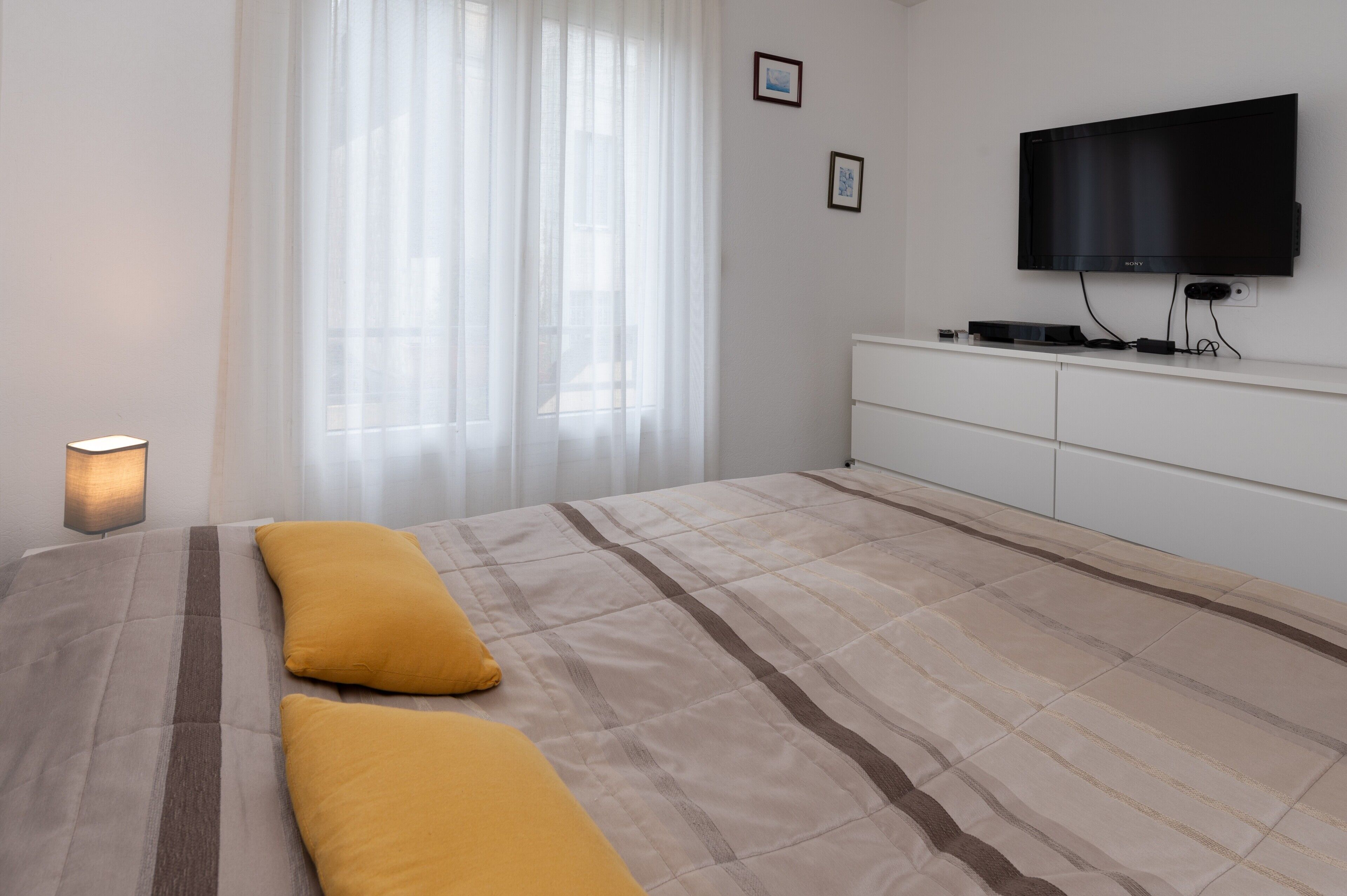 A bedroom with a bed, a TV mounted on the wall, a bedside table, and a window with curtains.