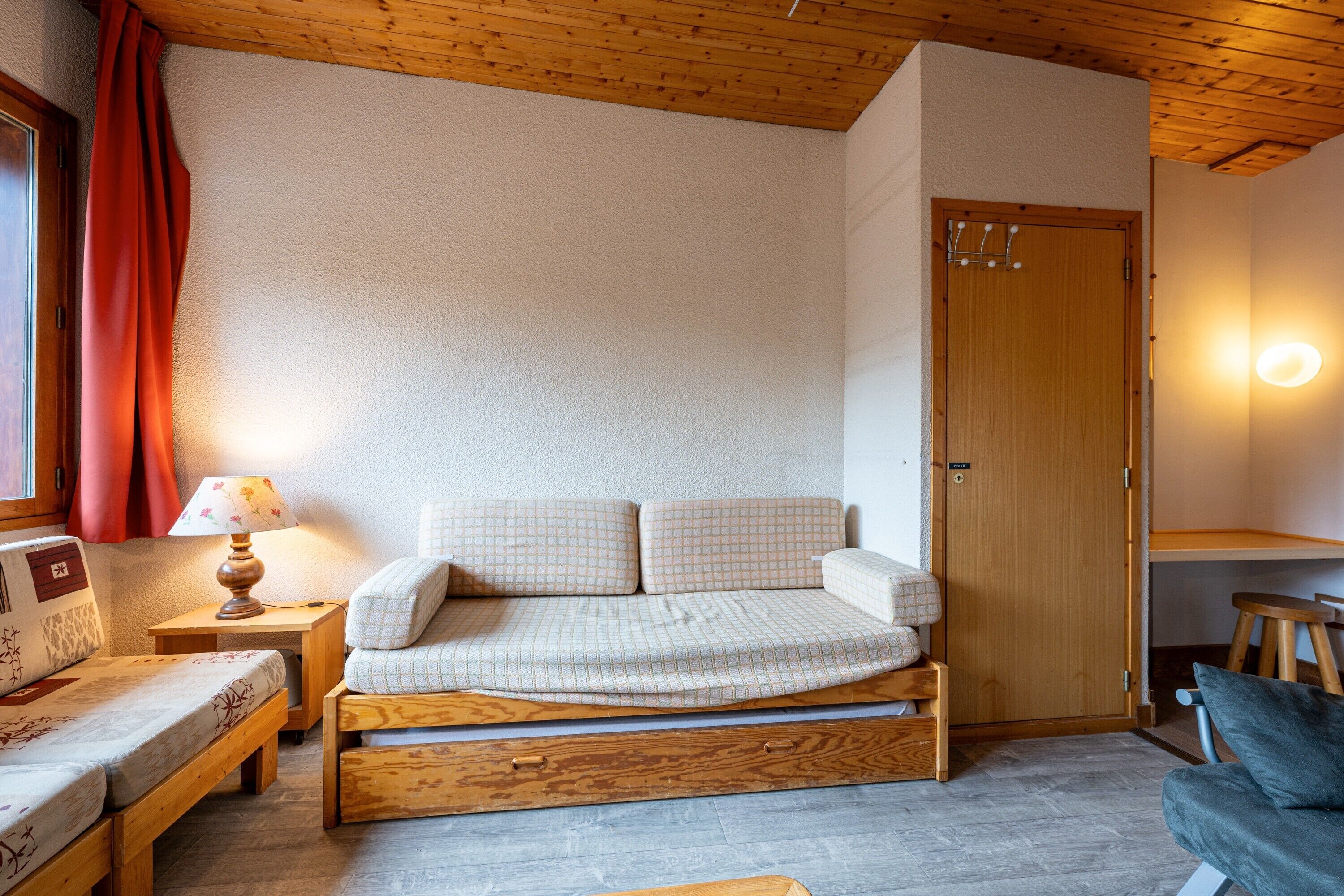 A cozy room with a bed, a wooden bench, a small table with a lamp, and a door.
