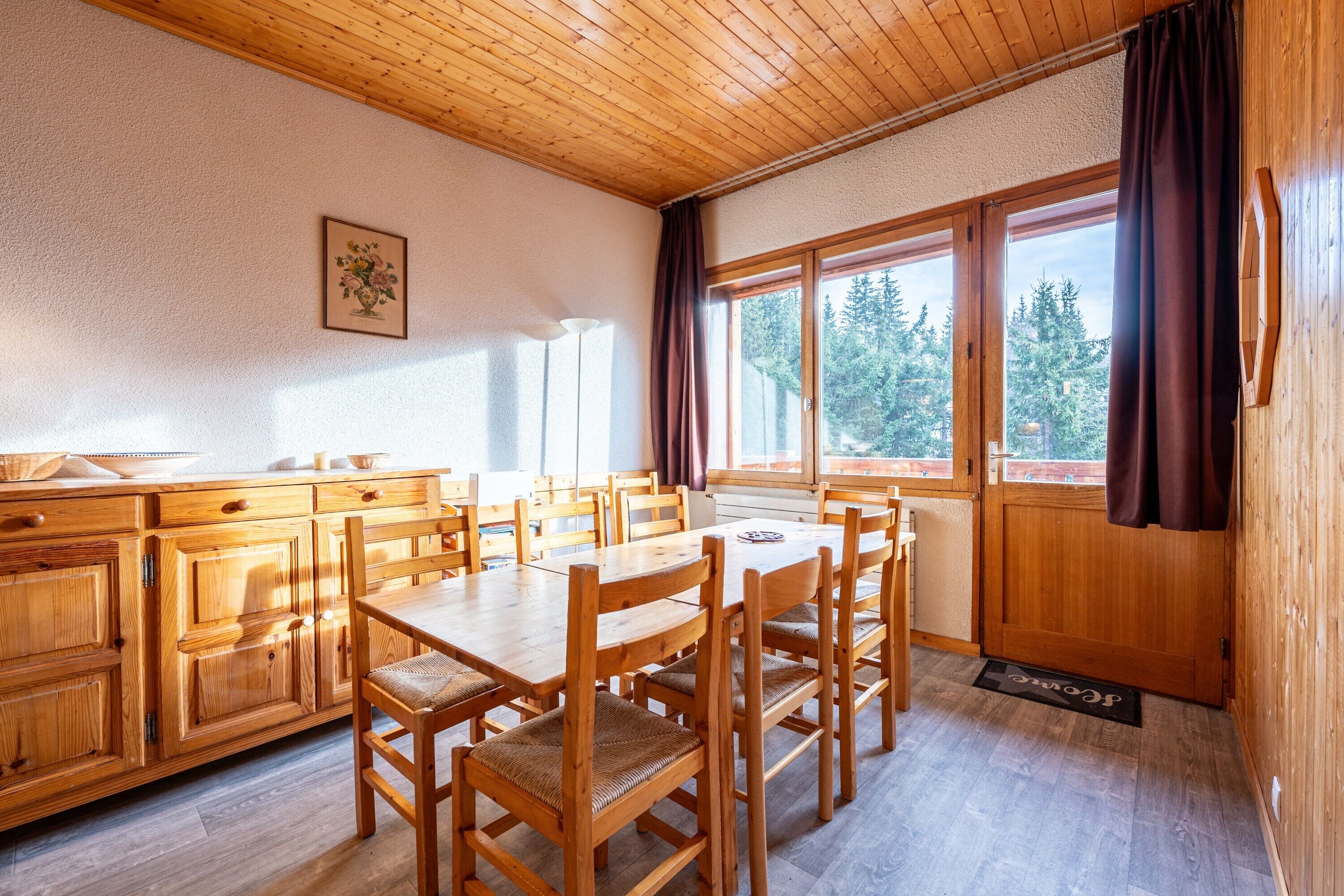 Comedor de madera con mesa y sillas, cocina con armarios de madera y ventanas grandes con cortinas.