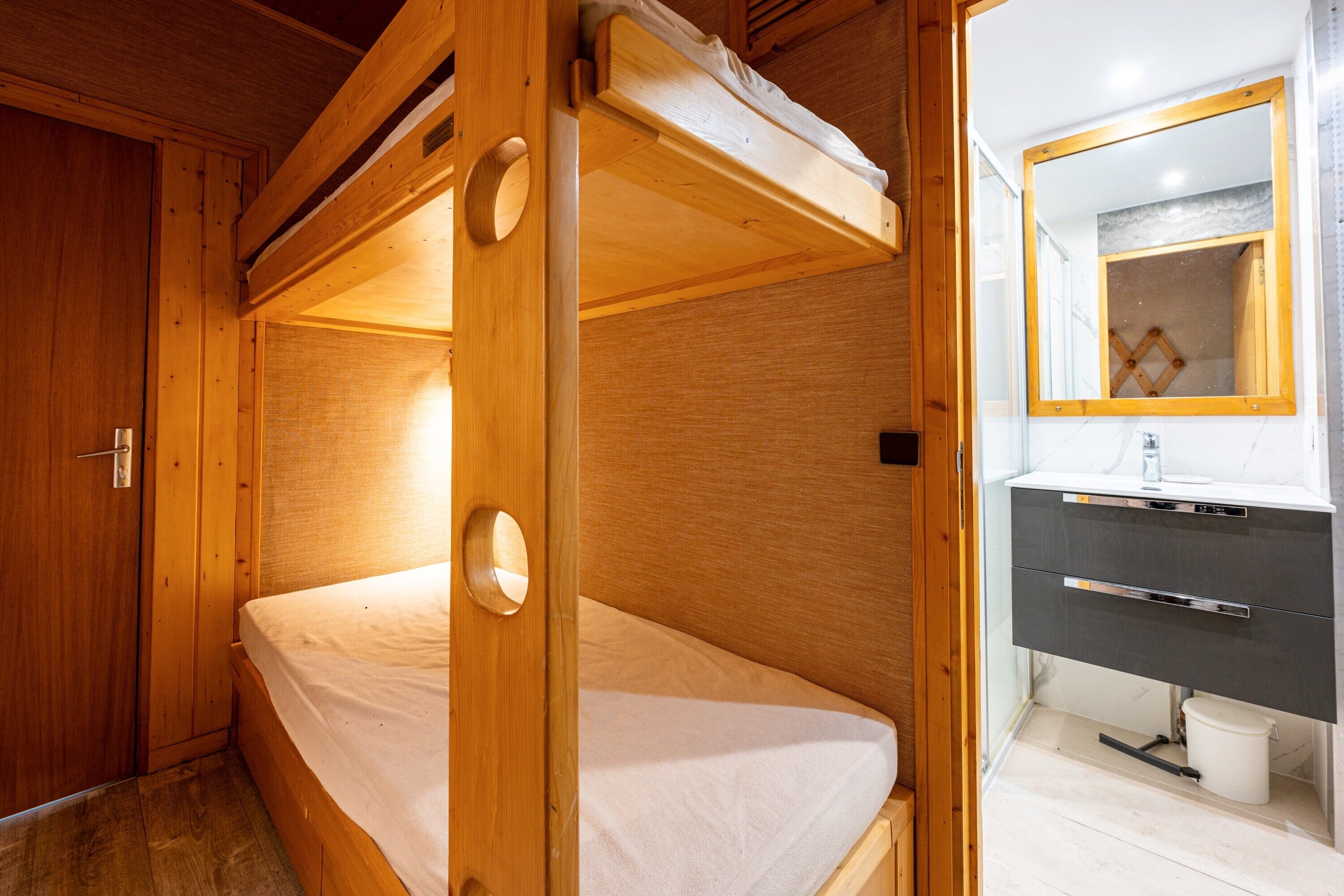 A bunk bed with a wooden frame, a bathroom with a mirror and sink, and a wooden door.