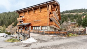 A wooden building with balconies, labeled 'TOUBKAL,' situated in a mountainous area with a parking lot and a snow-covered ground. - Apartment 26 – 9 Guests with Mountain View and Balcony (Les Allues)
