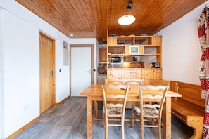 A wooden dining table with chairs, a wooden bench, a kitchen area with wooden cabinets, and a ceiling with wooden beams.