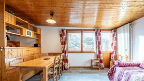 A wooden interior with a dining table, chairs, a bed with a purple quilt, and a window with curtains.