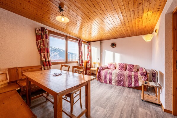 A wooden ceiling, a dining table with chairs, a sofa, a TV stand, and a window with curtains.