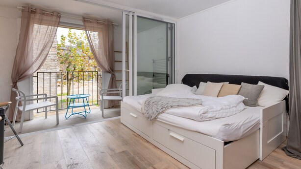 Un dormitorio con cama, una mesita, una silla y una puerta corrediza de vidrio que da a un balcón.