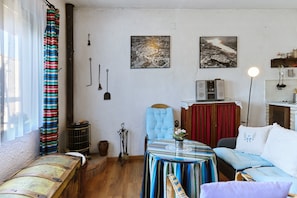 A cozy living room with a sofa, a table with a vase, and a chair. There are two paintings on the wall, a window with curtains, and a radiator.