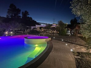 Une piscine à bord incurvé et dotée d’une bordure en pierre, entourée d’une allée pavée et d’un éclairage extérieur.