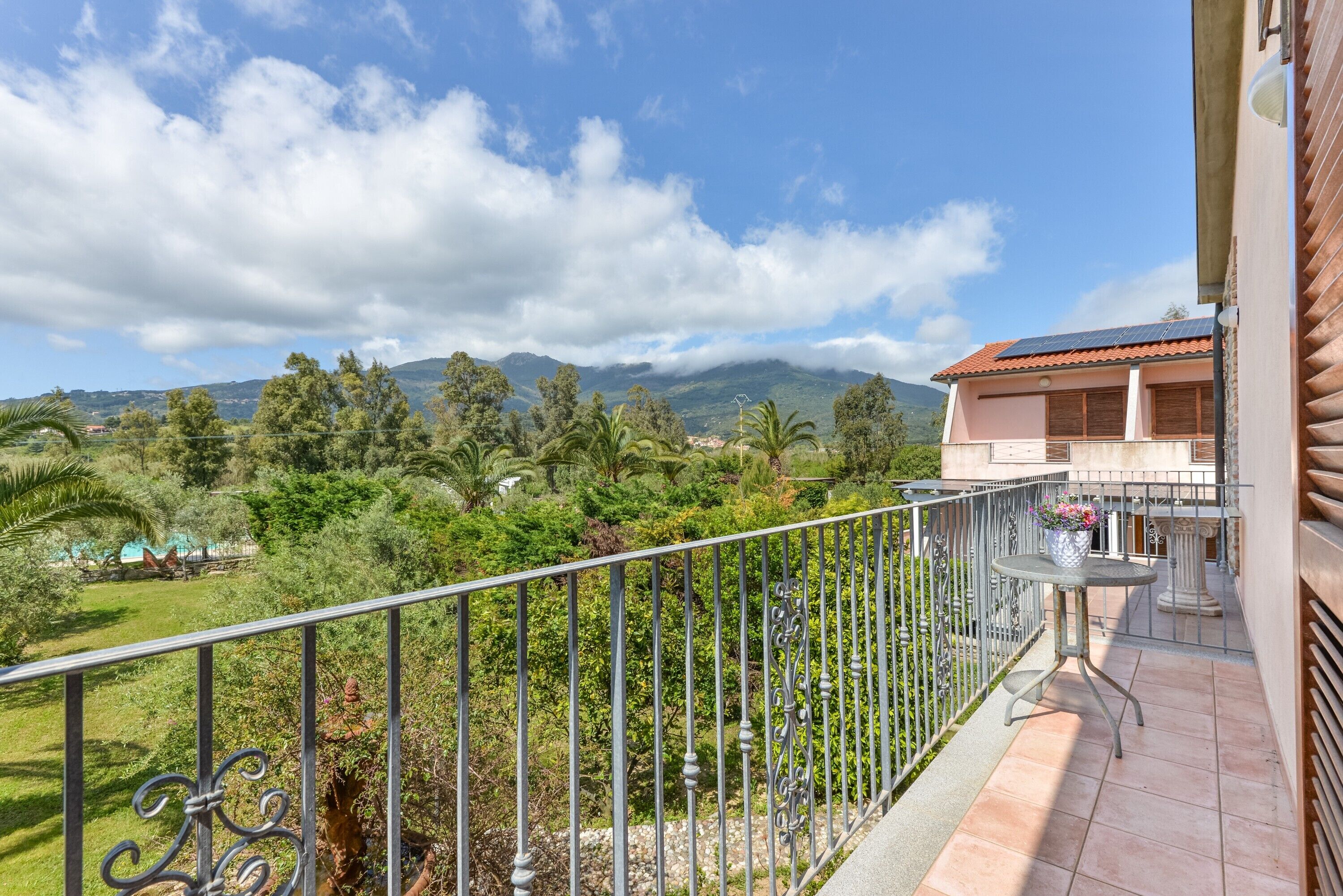 Ein Balkon mit Metallgeländer, ein kleiner Tisch mit Blumenvase und Blick auf eine gebirgige Landschaft.