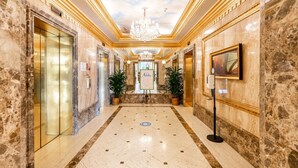 Un vestíbulo de hotel con pisos de mármol, ascensores, plantas en macetas, un candelabro y un mostrador de recepción.