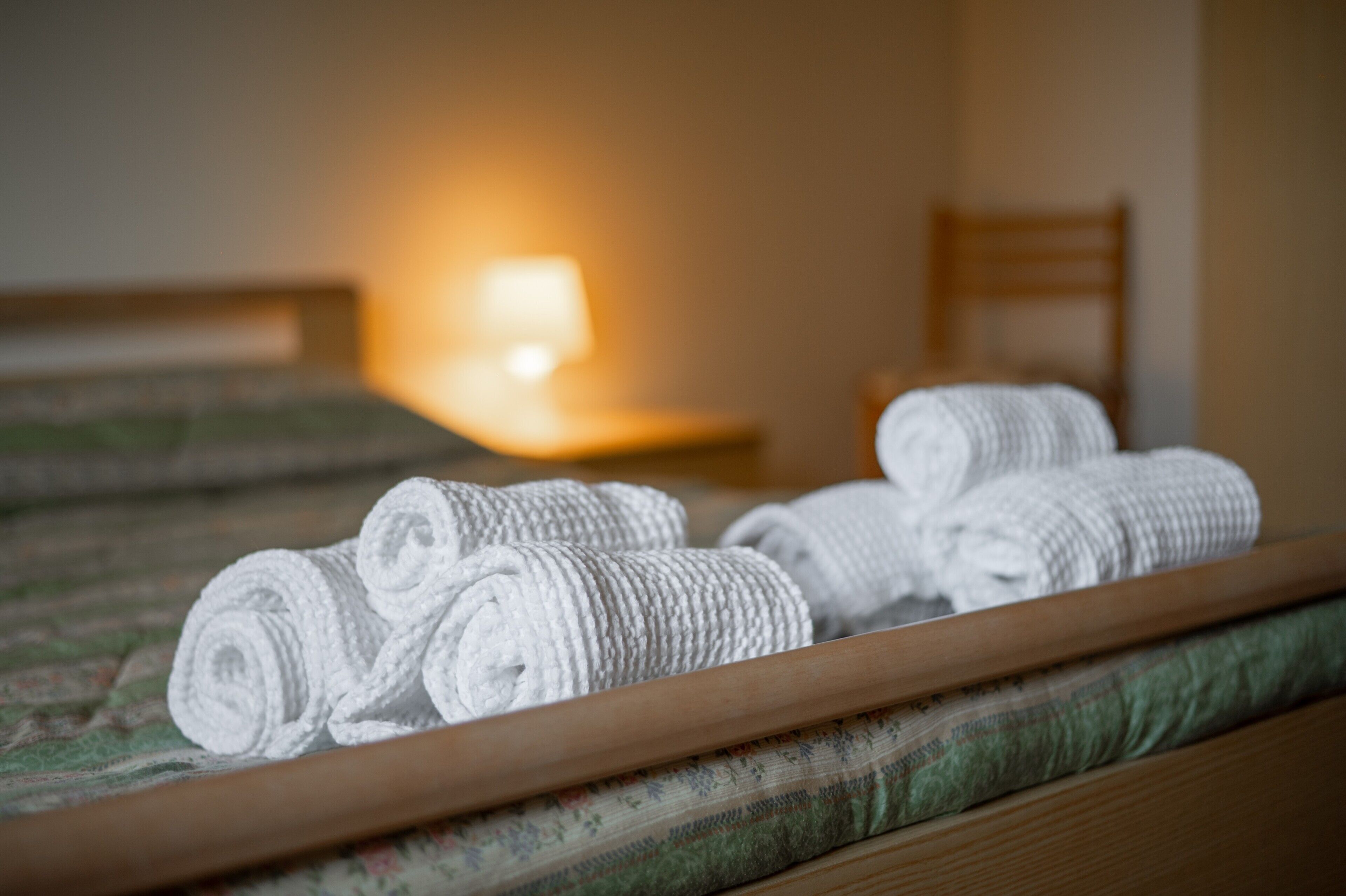 Un lit bien fait, avec des serviettes blanches pliées en rouleau, une lampe de chevet et une chaise en bois, dans une chambre d’hôtel.