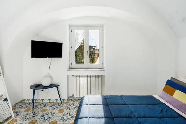 A bedroom with a bed, a television mounted on the wall, a small table with a lamp, and a window with a view of buildings outside.