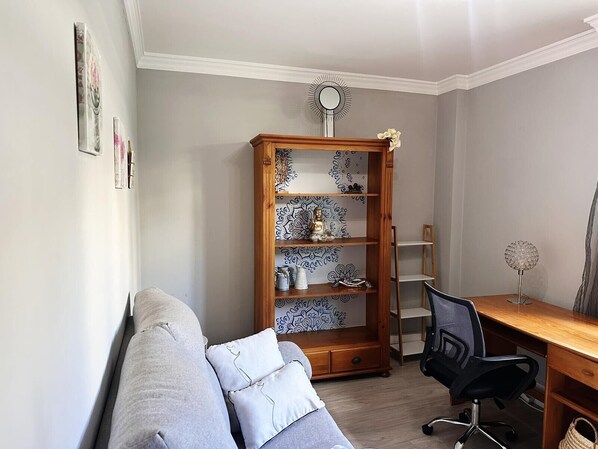 A room with a grey sofa, a wooden bookshelf, a desk with a chair, and a decorative mirror.