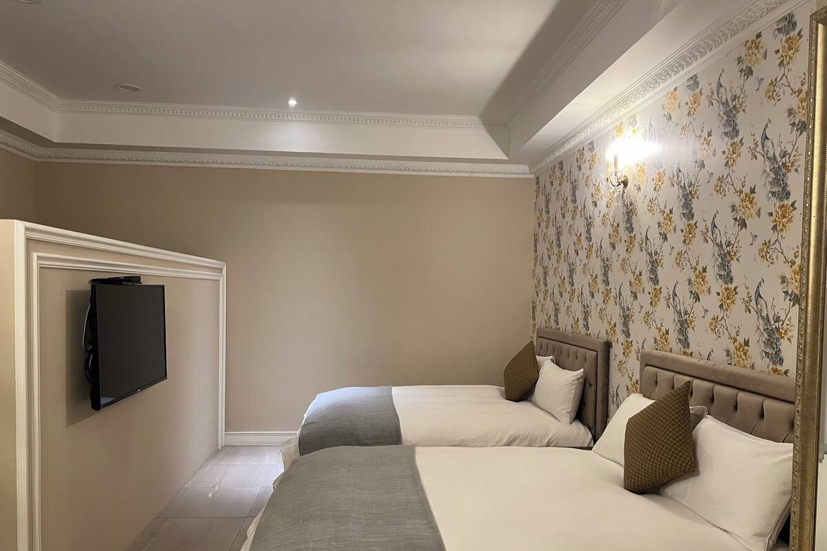 A hotel room with two beds, a flat-screen TV mounted on the wall, floral wallpaper, and a chandelier.