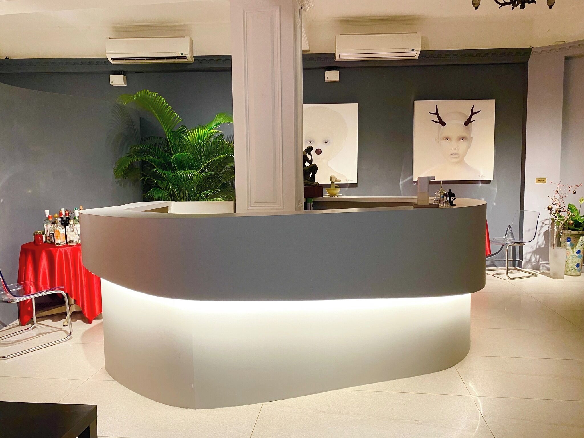 A reception area with a curved counter, air conditioning units, a red table with bottles, a plant, and artwork on the walls.