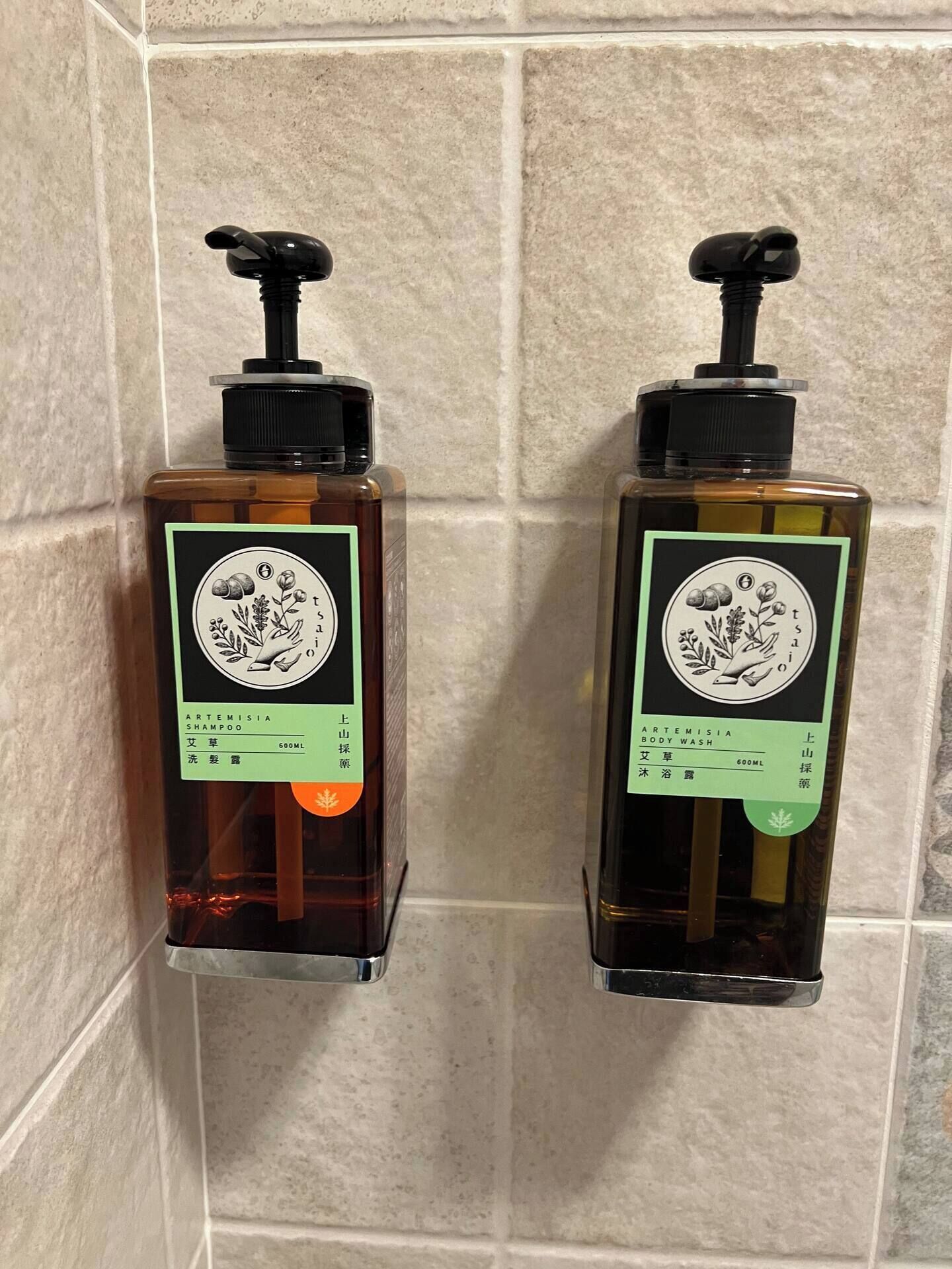Two bottles of shampoo and body wash on a tiled wall.