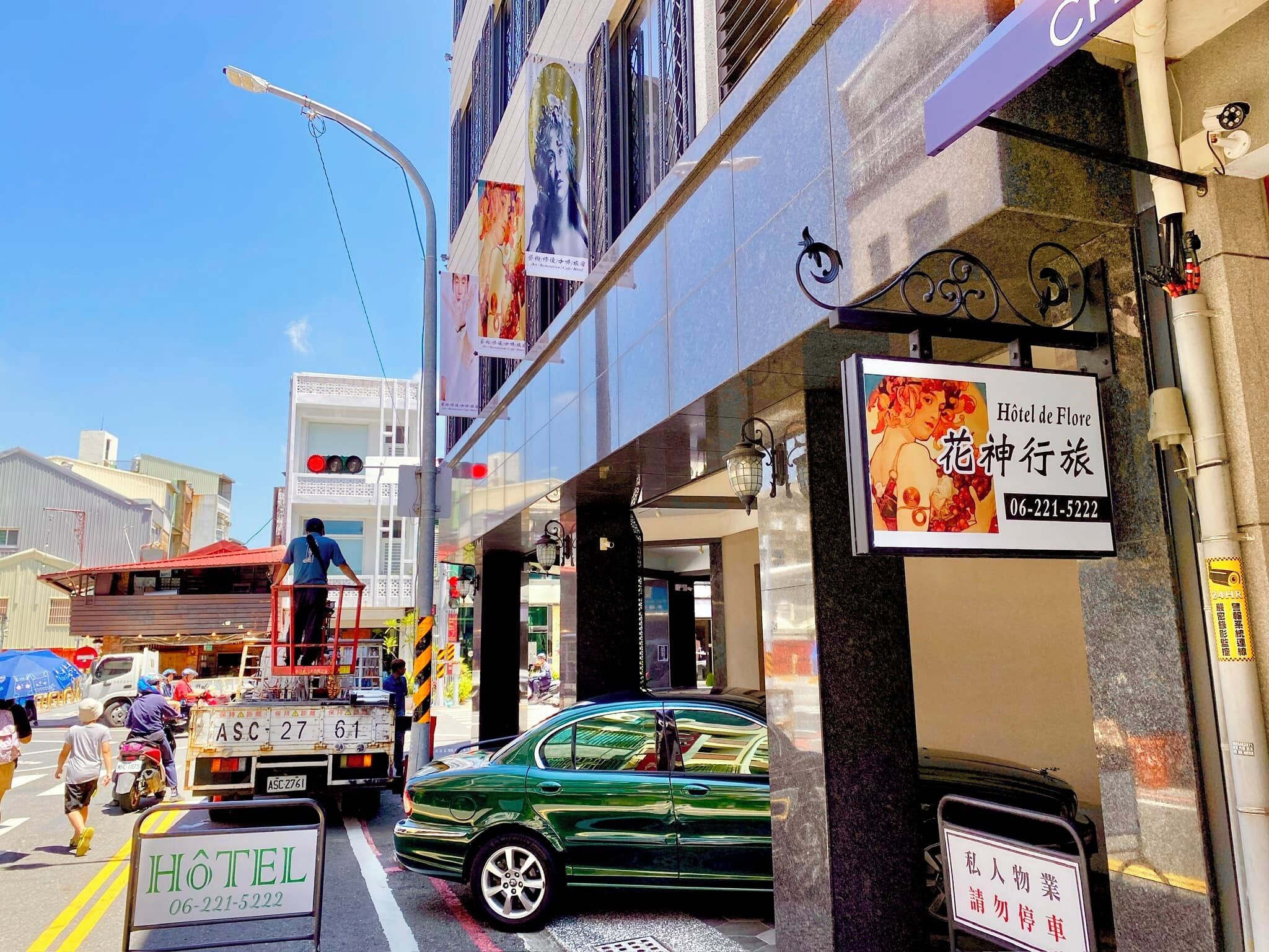 A street view with a hotel sign, a green car parked in front, and a building with a sign in a foreign language.