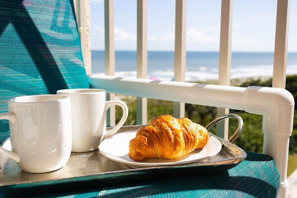 A breakfast setup with a croissant on a tray, two white mugs, and a view of the ocean.