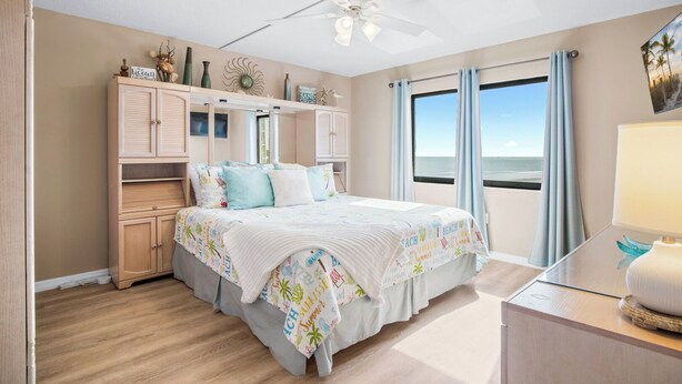 A bedroom with a bed, a ceiling fan, a nightstand, a window with curtains, and a view of the beach. - Beautiful Condo w/ Heated Pool & Stunning Ocean Views in Cape Canaveral! 🌊 (Cape Canaveral)