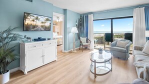 A spacious living room with a large window offering a view of the beach. There is a white cabinet, a round glass coffee table, and a blue sofa. - Beautiful Condo w/ Heated Pool & Stunning Ocean Views in Cape Canaveral! 🌊 (Cape Canaveral)
