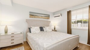 Une chambre à coucher comprenant un lit, des tables de chevet, une lampe, un tableau, un appareil de climatisation et une fenêtre avec des rideaux.