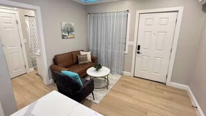 Una sala de estar con un sofá marrón, una mesa de centro redonda y una mesa blanca. Hay dos puertas blancas y una ventana con cortinas.