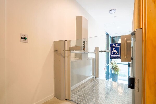 Entrada de un hotel con botón de ascensor, señal de acceso para sillas de ruedas y puerta de cristal.