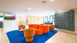 A modern hotel lobby with a blue carpet, orange seating, a TV on the wall, and a row of numbered lockers.