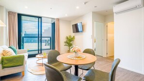 A modern living room with a round dining table, comfortable seating, a flat-screen TV, and a large window offering a view.