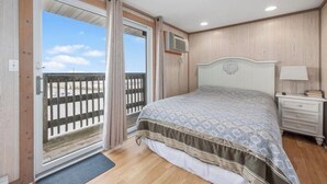 A bedroom with a bed, a nightstand, a lamp, a window with a view of a wooden deck, and a ceiling fan. - Charming 1-bedroom condo in tranquil Dennis with AC (Dennis)