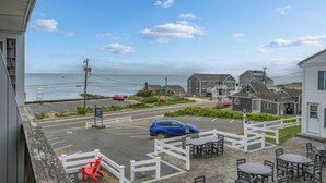 A coastal view with a parking area, a blue car, and a white fence with tables and chairs. - Charming 1-bedroom condo in tranquil Dennis with AC (Dennis)