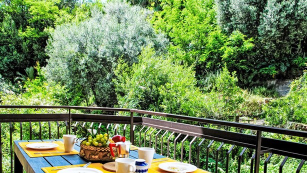 A wooden table set with plates, cups, and a bowl of fruit on a yellow mat, overlooking a lush garden. - La Conca by Interhome (Formia)