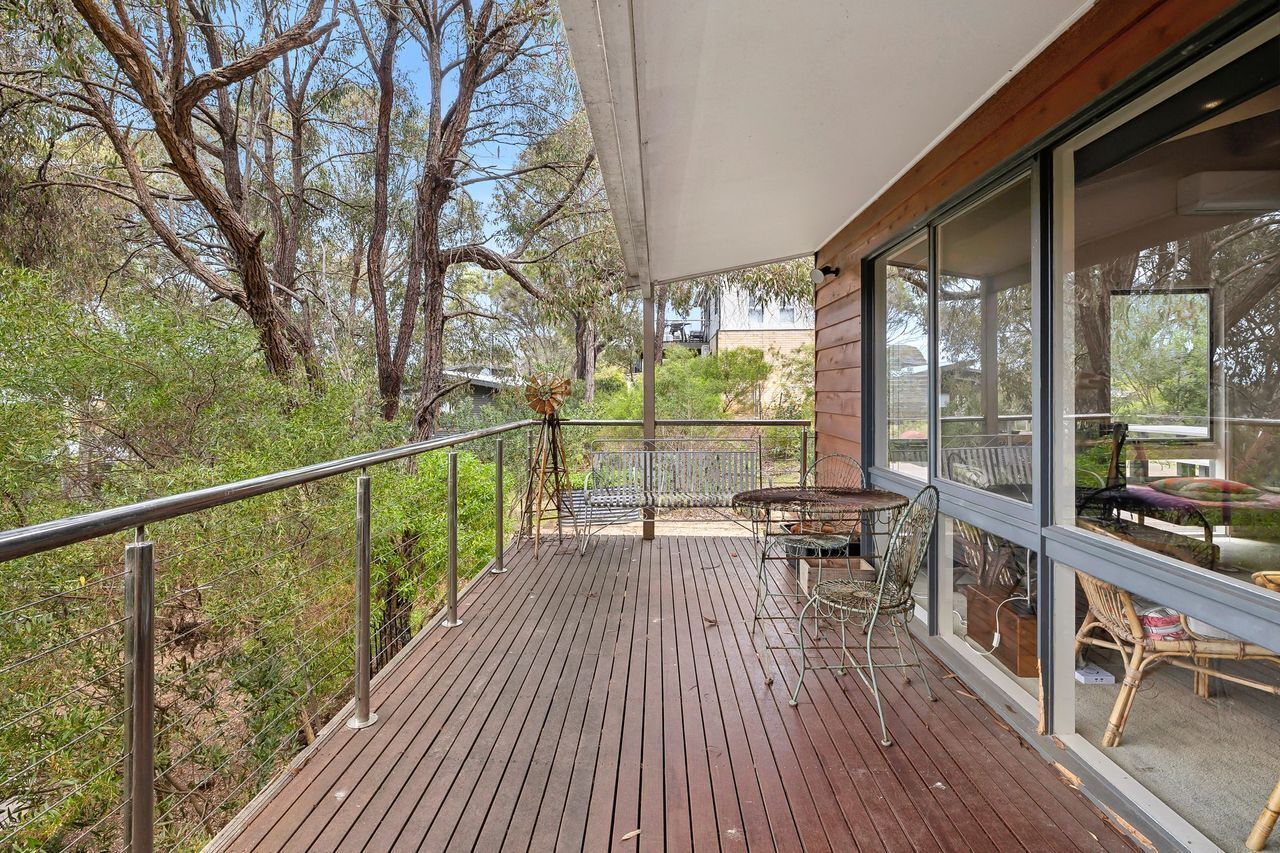 A wooden deck with metal railings, a table, and chairs, overlooking a natural landscape with trees and shrubs.