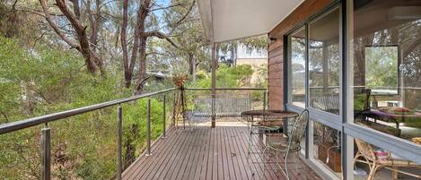 A wooden deck with metal railings, a table, and chairs, overlooking a natural landscape with trees and shrubs.