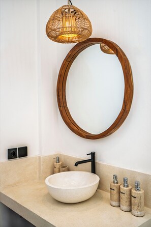 A bathroom with a round wooden-framed mirror, a white vessel sink, a black faucet, and three cylindrical bottles of toiletries. - Surga Villa by Betterplace (Canggu)