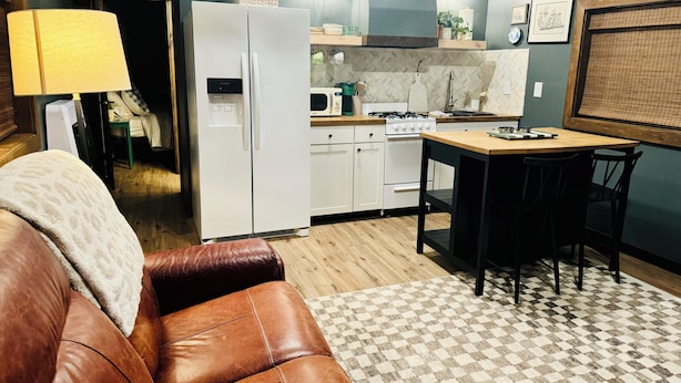 Un salon chaleureux avec un canapé en cuir marron, une table à manger avec des chaises, un coin cuisine doté de meubles blancs, un réfrigérateur et un four à micro-ondes.