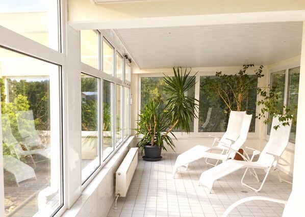 Un balcone baciato dal sole con sedie bianche, piante in vaso, ampie finestre e pavimento piastrellato.