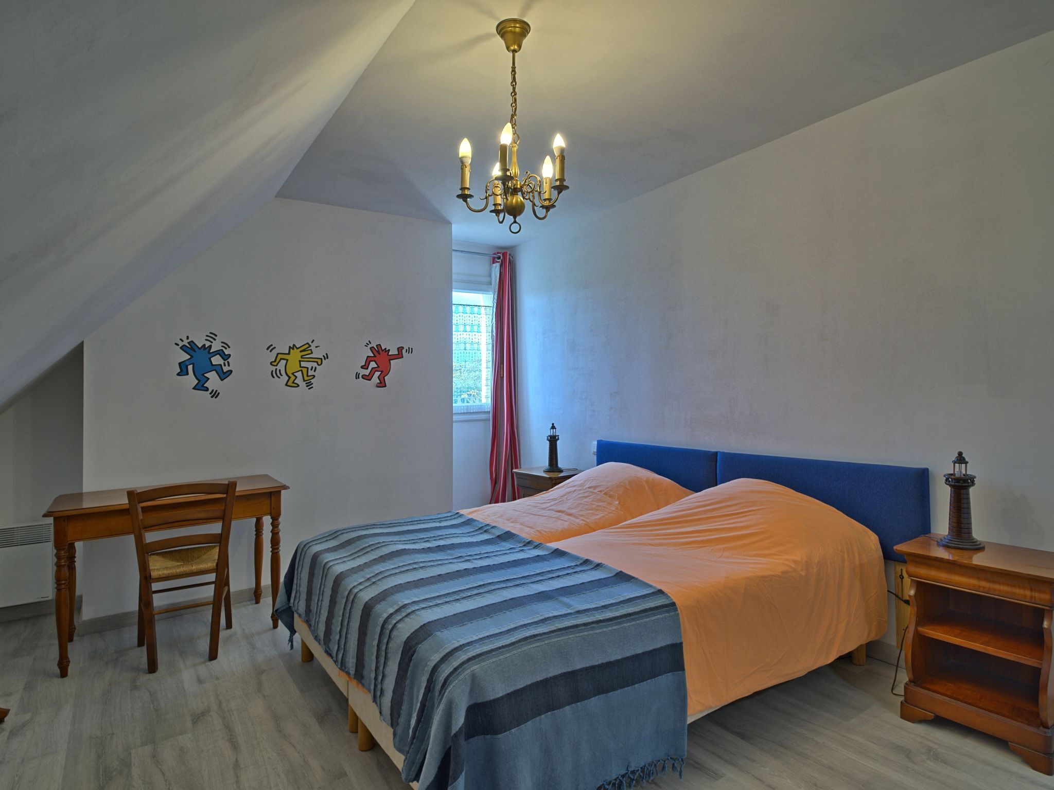 Un dormitorio con cama, escritorio y silla. Se observa una araña colgante sobre la cama y una mesita de noche con lámpara a un lado.