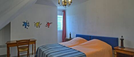 Un dormitorio con cama, escritorio y silla. Se observa una araña colgante sobre la cama y una mesita de noche con lámpara a un lado.