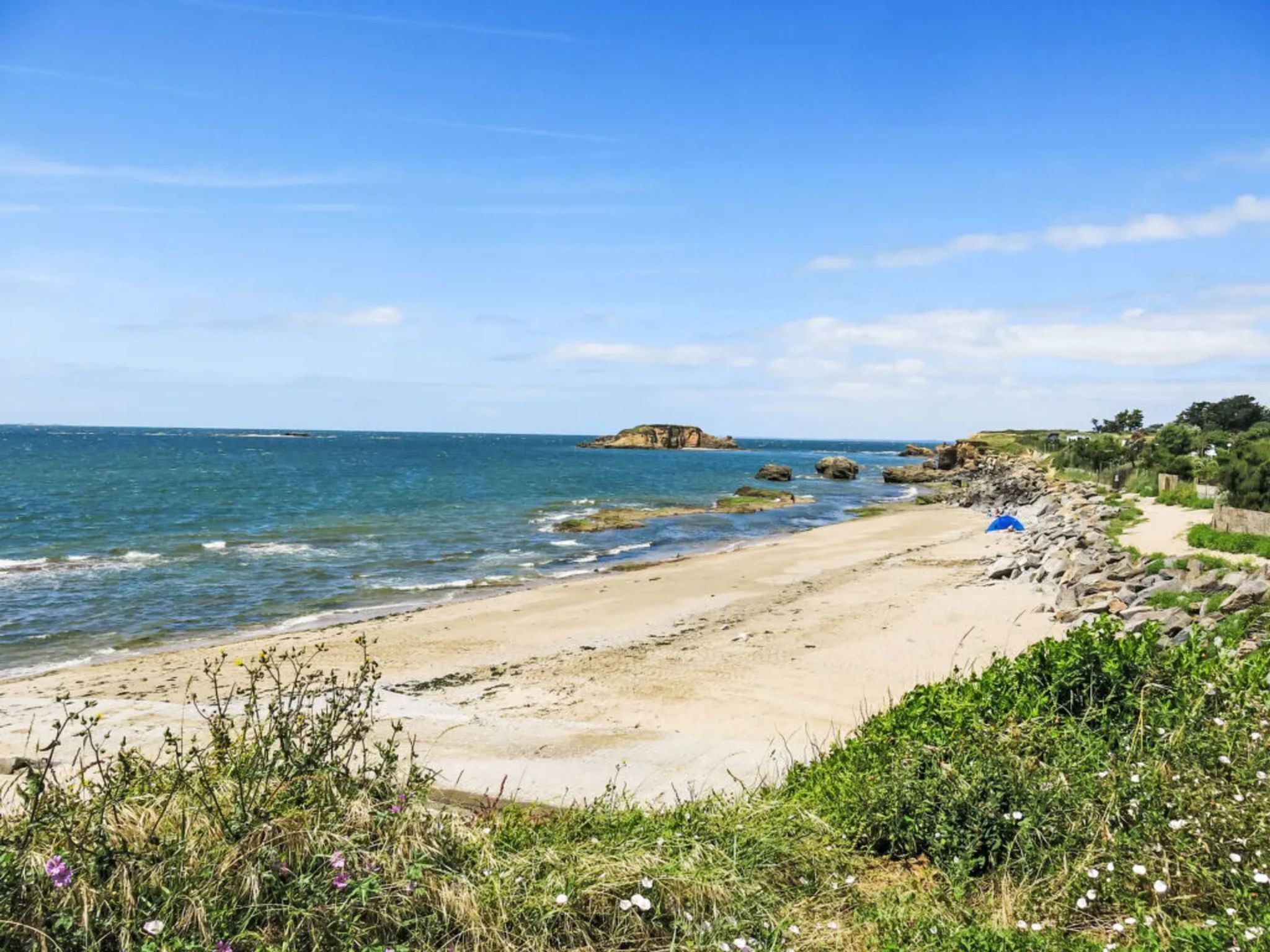 A coastal beach with clear skies, rocky outcrops, and a sandy path leading to a blue tent.