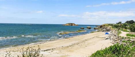 A coastal beach with clear skies, rocky outcrops, and a sandy path leading to a blue tent.