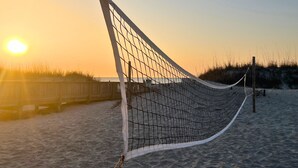 Ein Beachvolleyballnetz ist auf sandigem Strand aufgebaut, im Hintergrund ist ein Holzzaun zu sehen und in der Ferne geht die Sonne unter.