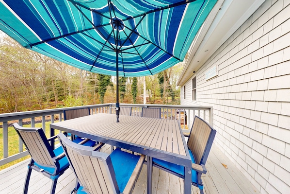 Relax on the deck surrounded by a serene wooded environment, with a table and chairs under a shaded umbrella.