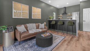 Ein modernes Wohnzimmer mit einer Couch, einem Hocker und Barhocker. Es gibt eine Küchenzeile mit einem Kühlschrank und einem Fenster.