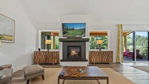 A spacious living room with a fireplace, a television mounted above it, and a large window offering a view of the outdoors. - Idaho Club Golf Course | Luxe Living & Hot Tub (Sandpoint)