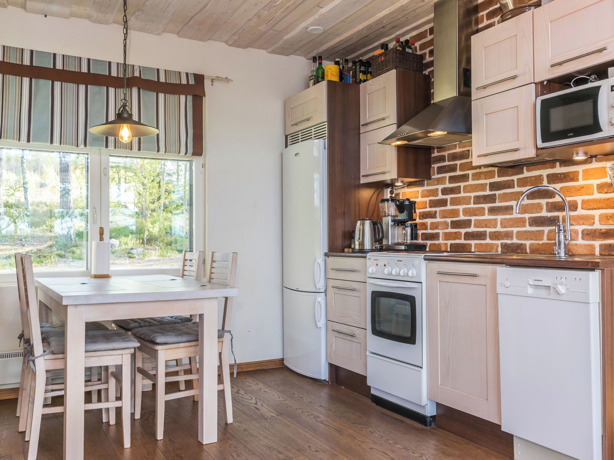 A kitchen with a brick wall, white cabinets, a stainless steel range hood, a built-in microwave, and a dining area with a table and chairs.