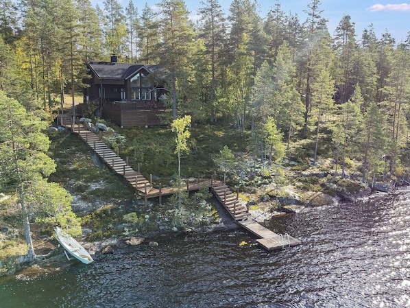 Una cabaña con terraza de madera que da a un muelle sobre el agua, rodeada de un bosque denso.