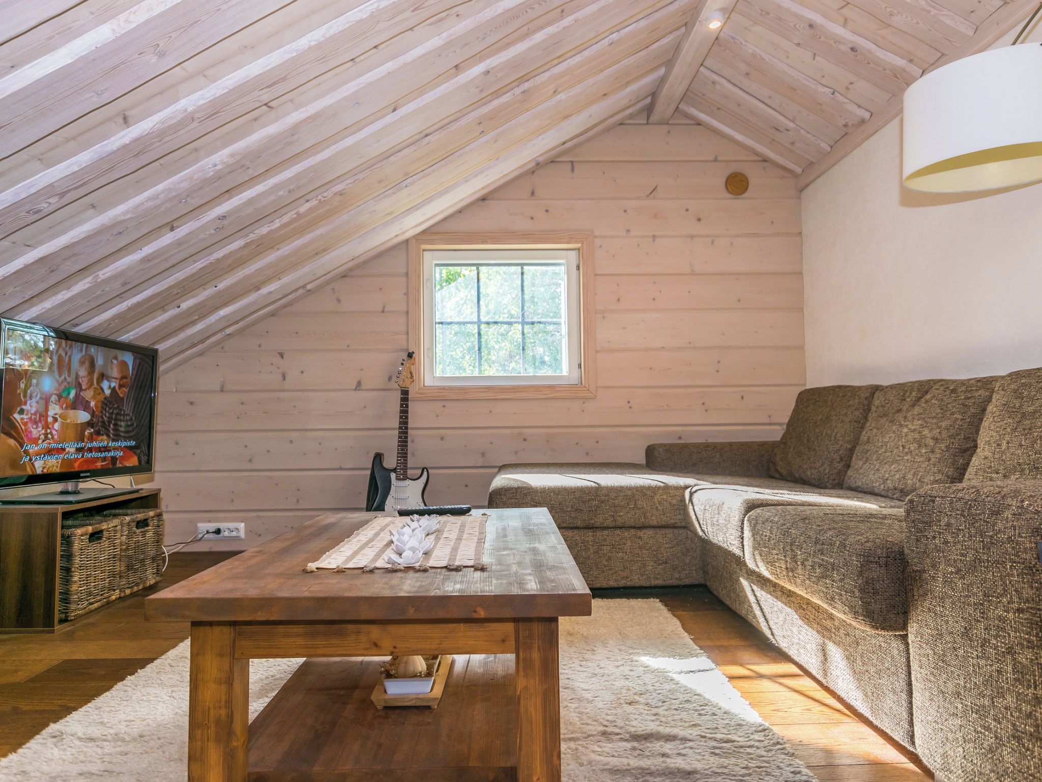 Quarto de loft aconchegante com teto inclinado, uma janela grande, televisão, um violão, sofá e mesa de madeira.