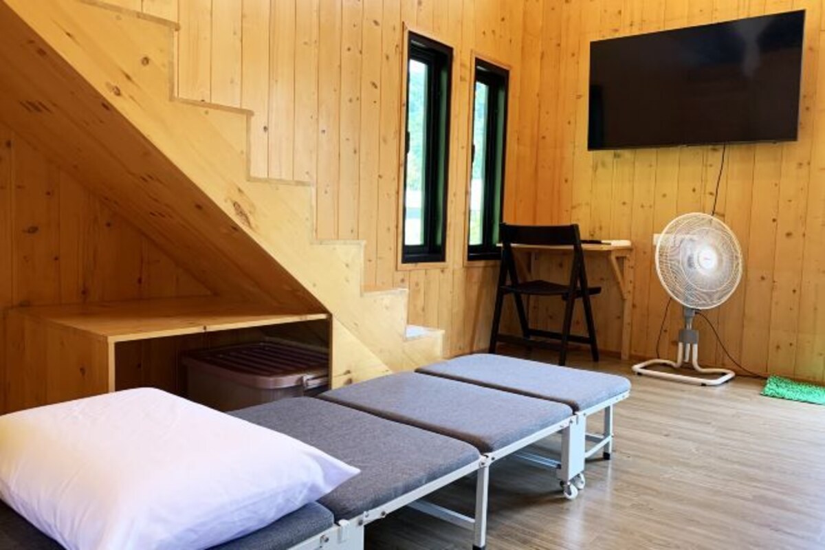 Una cabaña con interior de madera, una cama con almohada blanca, un banco, un ventilador y un televisor montado en la pared.